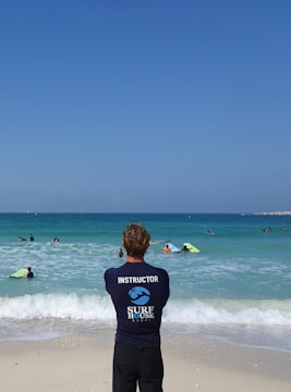 A surf instructor stands on the sandy beach with their back to the camera, observing a group of people in the ocean. Some individuals are using bodyboards on the waves. The sky is clear and blue, while the sea exhibits various shades of blue and turquoise.