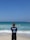A surf instructor stands on the sandy beach with their back to the camera, observing a group of people in the ocean. Some individuals are using bodyboards on the waves. The sky is clear and blue, while the sea exhibits various shades of blue and turquoise.