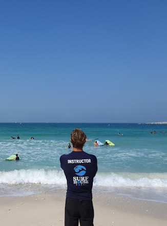 A surf instructor stands on the sandy beach with their back to the camera, observing a group of people in the ocean. Some individuals are using bodyboards on the waves. The sky is clear and blue, while the sea exhibits various shades of blue and turquoise.