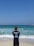 A surf instructor stands on the sandy beach with their back to the camera, observing a group of people in the ocean. Some individuals are using bodyboards on the waves. The sky is clear and blue, while the sea exhibits various shades of blue and turquoise.