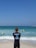 A surf instructor stands on the sandy beach with their back to the camera, observing a group of people in the ocean. Some individuals are using bodyboards on the waves. The sky is clear and blue, while the sea exhibits various shades of blue and turquoise.