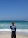 A surf instructor stands on the sandy beach with their back to the camera, observing a group of people in the ocean. Some individuals are using bodyboards on the waves. The sky is clear and blue, while the sea exhibits various shades of blue and turquoise.