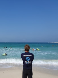 A surf instructor stands on the sandy beach with their back to the camera, observing a group of people in the ocean. Some individuals are using bodyboards on the waves. The sky is clear and blue, while the sea exhibits various shades of blue and turquoise.