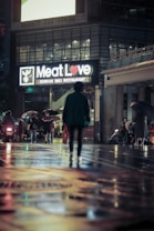 A dimly lit street scene at night featuring a brightly lit sign for a Korean BBQ restaurant called 'Meat Love.' The street is wet, possibly from rain, and reflects colorful lights. A person walks alone towards the restaurant, carrying a bag. Several other people are visible in the background, some with umbrellas, and there are a few vehicles parked alongside.