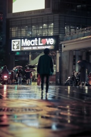 A dimly lit street scene at night featuring a brightly lit sign for a Korean BBQ restaurant called 'Meat Love.' The street is wet, possibly from rain, and reflects colorful lights. A person walks alone towards the restaurant, carrying a bag. Several other people are visible in the background, some with umbrellas, and there are a few vehicles parked alongside.
