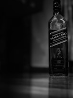 A bottle of Johnnie Walker Black Label blended scotch whisky stands on a polished surface. The image is monochromatic, creating a moody and elegant atmosphere.