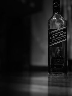 A bottle of Johnnie Walker Black Label blended scotch whisky stands on a polished surface. The image is monochromatic, creating a moody and elegant atmosphere.
