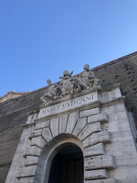 A stone entrance with a grand archway leading into a building, adorned with classical sculptures. Above the archway, carved statues depict several figures in traditional garb, and the words 'MVSEI VATICANI' are engraved below them. The structure features detailed masonry with large, rectangular blocks. The sky is clear blue, providing a serene backdrop.