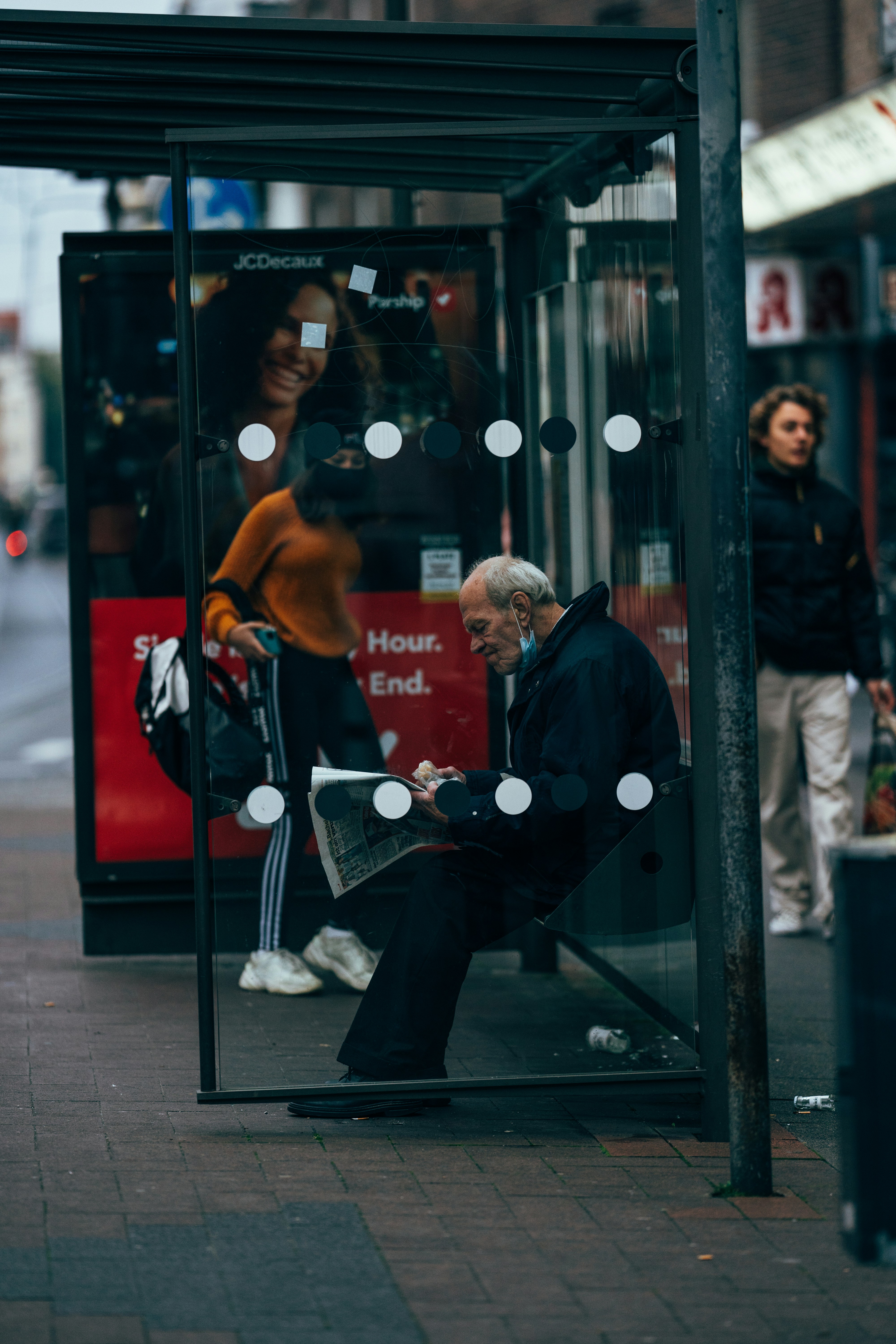 Woman reading newspaper alone at bus stop, pigeons around her feet, morning light
