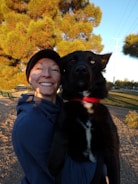 A person wearing a blue hoodie and a black beanie is holding a large black and white dog. They are in an outdoor setting with lush green pine trees in the background, and the scene is lit by warm natural sunlight.