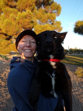 A person wearing a blue hoodie and a black beanie is holding a large black and white dog. They are in an outdoor setting with lush green pine trees in the background, and the scene is lit by warm natural sunlight.