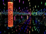 A dark room filled with an array of suspended vertical light tubes, each glowing in vibrant colors like orange, blue, green, and pink. The reflective surfaces create an effect of infinite lights and colors, giving a sense of depth and immersion. A few people can be seen observing the installation, adding a human element to the scene.