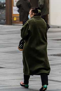 A person wearing a long green coat and a face mask is walking on a paved street. They are carrying a black handbag with a colorful pattern.
