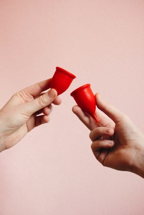 Two hands are holding red menstrual cups against a soft pink background. The hands are positioned to face each other, with each hand holding a single cup between the thumb and fingers.