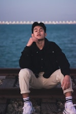 A person sits on railroad tracks near a body of water. The individual appears relaxed, wearing casual attire including a black jacket, light pants, striped socks, and white sneakers. The background features a calm expanse of water with lights visible on the far shore.