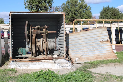 A metal enclosure appears to be housing a large industrial mechanism with gears and a wound cable. The enclosure is partially open, revealing rust and wear on the equipment. Behind the structure, there are trees and a sidewalk, indicating an outdoor setting. The ground is covered with grass, and there's a pathway leading to the setup.