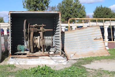 A metal enclosure appears to be housing a large industrial mechanism with gears and a wound cable. The enclosure is partially open, revealing rust and wear on the equipment. Behind the structure, there are trees and a sidewalk, indicating an outdoor setting. The ground is covered with grass, and there's a pathway leading to the setup.