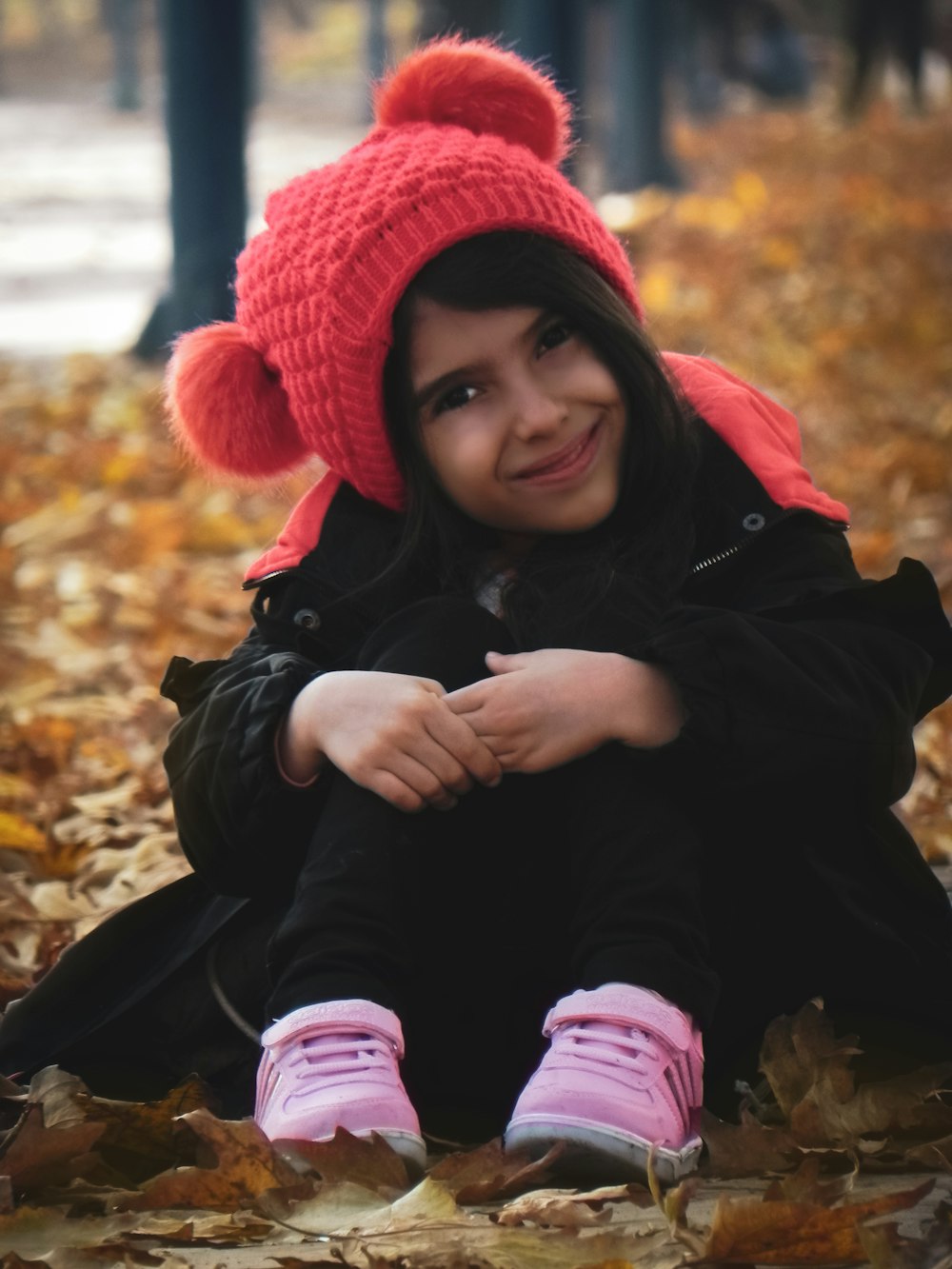 girl in black jacket and red knit cap sitting on dried leaves during daytime