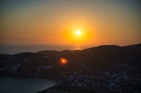 A serene landscape featuring a large, bright sun setting over a calm sea. The sky is awash with warm gradients of orange and yellow, casting a gentle light over distant hills and a coastal village below.
