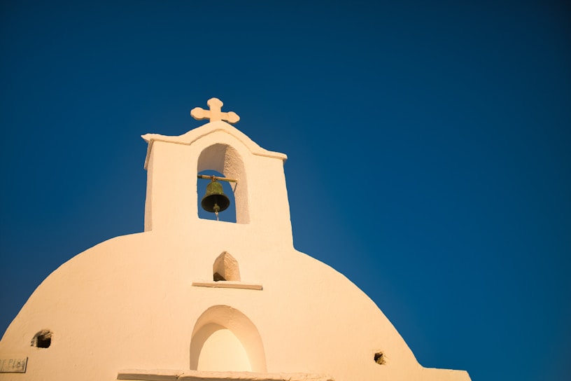 A white church with a rounded top features a small bell tower and a prominent cross against a clear blue sky. The architecture is simple yet elegant, highlighting Mediterranean influence.