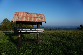 A wooden structure with a tiled roof stands in a grassy field with a sign containing text in Croatian. The background reveals a panoramic view of a vast landscape with scattered trees under a clear blue sky.