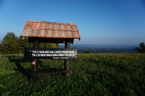 A wooden structure with a tiled roof stands in a grassy field with a sign containing text in Croatian. The background reveals a panoramic view of a vast landscape with scattered trees under a clear blue sky.