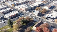 An aerial view of a town intersection showing multiple buildings, parking lots, roads with cars, and a park area with trees in various shades of green, yellow, and red.
