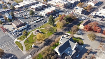 Aerial view of a small town featuring a central park with green spaces, intersected by multiple streets and a railroad track. The streets are lined with various commercial buildings, and there are parked vehicles in multiple parking lots. Trees with autumn foliage add color to the scene.