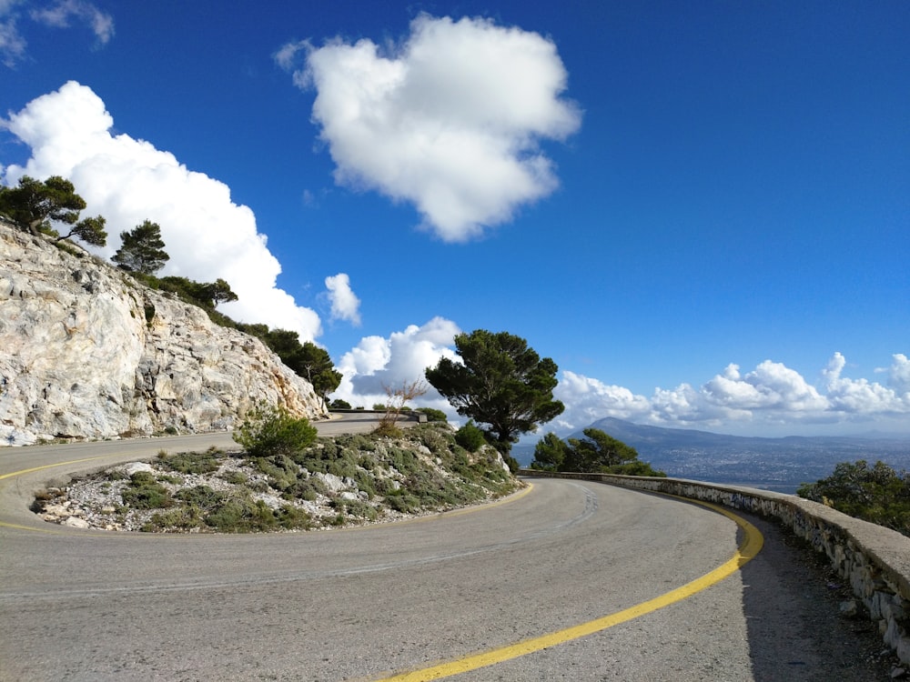 Graue Betonstraße in der Nähe grüner Bäume unter blauem Himmel und weißen Wolken tagsüber