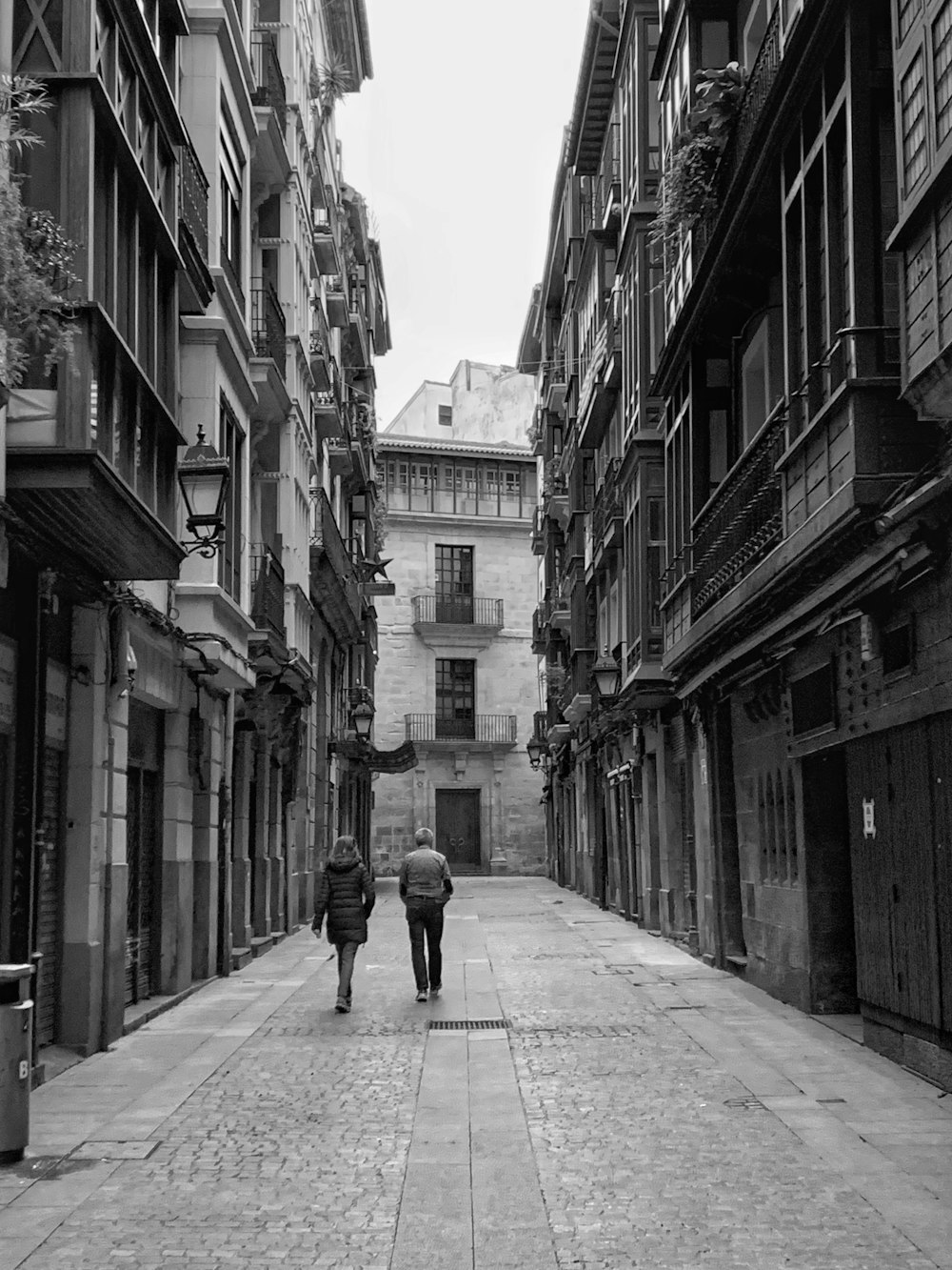 grayscale photo of woman walking on sidewalk between buildings