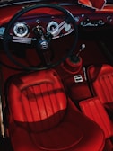 The interior of a vintage car features a sleek dashboard with circular gauges and a classic steering wheel. The seats and surrounding trim are covered in rich red leather, complementing the elegant design.