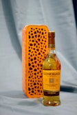 A bottle of Glenmorangie The Original Single Malt Scotch Whisky is placed in front of an orange box with a decorative pattern. The bottle has a golden hue, indicating the rich color of the whisky. The setting appears to be simple with a soft blue fabric as a backdrop.