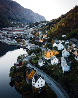 A scenic town nestled in a valley beside a body of water, featuring colorful houses with steep rooftops lining winding roads. The landscape is lush with trees, transitioning to mountainous terrain in the background. The architecture stands out against the earthy tones of the natural surroundings.