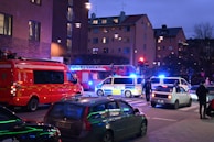 A nighttime street scene with emergency response vehicles, including police vans and a fire truck, parked at an intersection. Blue and red lights illuminate the area as pedestrians and other cars are present. The backdrop consists of residential buildings with glowing windows, indicating an urban setting.