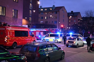 A nighttime street scene with emergency response vehicles, including police vans and a fire truck, parked at an intersection. Blue and red lights illuminate the area as pedestrians and other cars are present. The backdrop consists of residential buildings with glowing windows, indicating an urban setting.