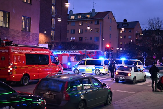 A nighttime street scene with emergency response vehicles, including police vans and a fire truck, parked at an intersection. Blue and red lights illuminate the area as pedestrians and other cars are present. The backdrop consists of residential buildings with glowing windows, indicating an urban setting.