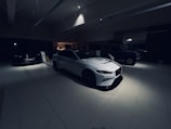 A dimly lit car showroom with multiple vehicles on display, featuring a white sports car prominently in the foreground. The setting is modern and sleek, with clean lines and minimalist design. The lighting is focused on the white car, creating a dramatic contrast with the darker surroundings. Other vehicles are partially visible in the background.