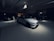 A dimly lit car showroom with multiple vehicles on display, featuring a white sports car prominently in the foreground. The setting is modern and sleek, with clean lines and minimalist design. The lighting is focused on the white car, creating a dramatic contrast with the darker surroundings. Other vehicles are partially visible in the background.