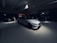 A dimly lit car showroom with multiple vehicles on display, featuring a white sports car prominently in the foreground. The setting is modern and sleek, with clean lines and minimalist design. The lighting is focused on the white car, creating a dramatic contrast with the darker surroundings. Other vehicles are partially visible in the background.