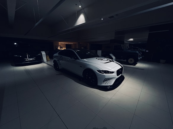 A dimly lit car showroom with multiple vehicles on display, featuring a white sports car prominently in the foreground. The setting is modern and sleek, with clean lines and minimalist design. The lighting is focused on the white car, creating a dramatic contrast with the darker surroundings. Other vehicles are partially visible in the background.