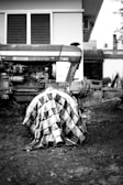 A tractor parked beside a building with a metal sheet draped over one of its large wheels. The tractor is positioned on rough terrain, and the image is in black and white, highlighting the details of the tractor's engine and the texture of the metal sheet.