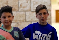 Two boys are standing side by side, both looking at the camera. They are casually dressed; the boy on the left wears a dark-colored shirt with a turquoise collar and pink accent, while the boy on the right wears a blue sweatshirt with a white logo. The background is a stone wall with a few blurred details.