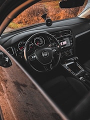 The interior of a car showing the steering wheel with a car brand logo, instrument panel displaying speedometer and tachometer, gear shift, touchscreen navigation system, and dashboard. The road outside is visible through the windshield, bordered by dry grass.