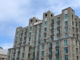 A multi-story hotel building with a modern architectural design featuring numerous windows and balconies. The structure is made of gray stone, and the word 'HOTEL' is prominently displayed at the top. The sky is partly cloudy, creating a peaceful atmosphere.