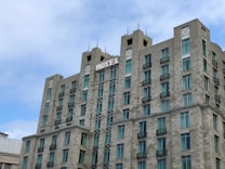 A multi-story hotel building with a modern architectural design featuring numerous windows and balconies. The structure is made of gray stone, and the word 'HOTEL' is prominently displayed at the top. The sky is partly cloudy, creating a peaceful atmosphere.