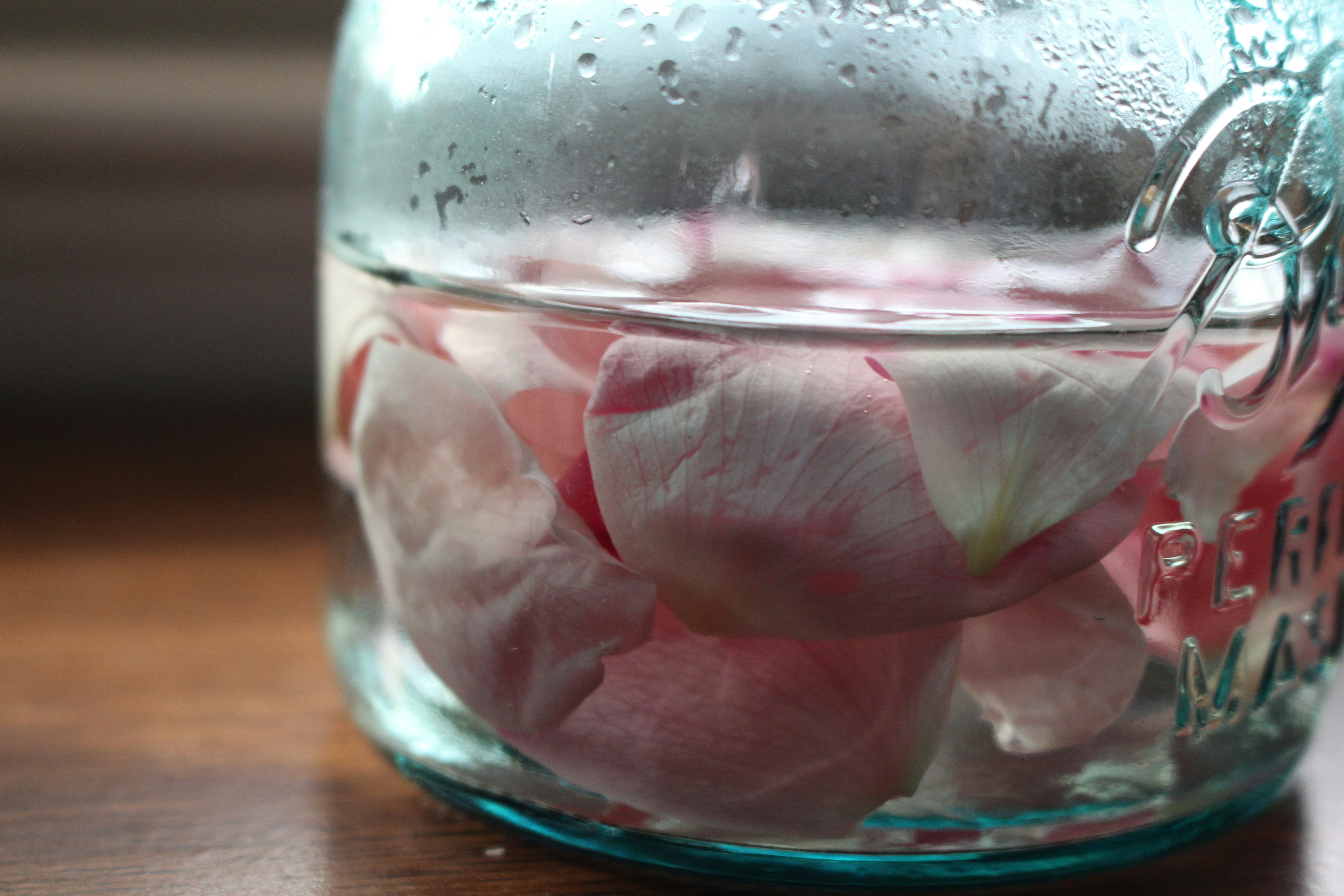 Clear glass jar of homemade rosewater next to fresh rose petals - Benefits of rosewater