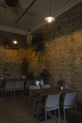 An inviting interior space with exposed brick walls and ambient lighting. Several potted plants add a touch of greenery. The room features modern chairs and wooden tables arranged for dining, with a minimalist and cozy aesthetic.