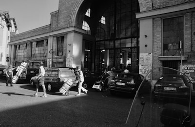 Several people are moving trolleys loaded with pallets across a street. The scene is set in front of a large building with arched windows, possibly an urban market or warehouse. Cars are parked along the street, and the area is bustling with activity.