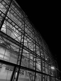 A detailed view of a modern architectural structure with a glass facade showcasing intricate steel beams and structural components. The image is in black and white, highlighting the contrast between the illuminated interior and the dark exterior.