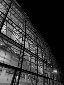 A detailed view of a modern architectural structure with a glass facade showcasing intricate steel beams and structural components. The image is in black and white, highlighting the contrast between the illuminated interior and the dark exterior.